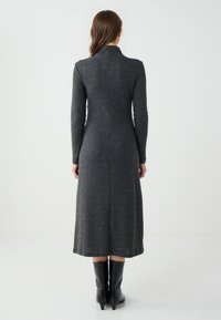 Langes graues Kleid aus strukturiertem Strickstoff, mit hohem Kragen und langen Ärmeln, das unterhalb des Knies endet, kombiniert mit schwarzen Stiefeln.