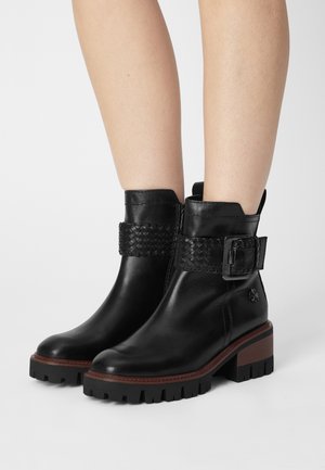 Botas de cuero negro hasta el tobillo con una correa trenzada y detalle de hebilla, que presentan un tacón grueso y suela de goma con un patrón texturizado.