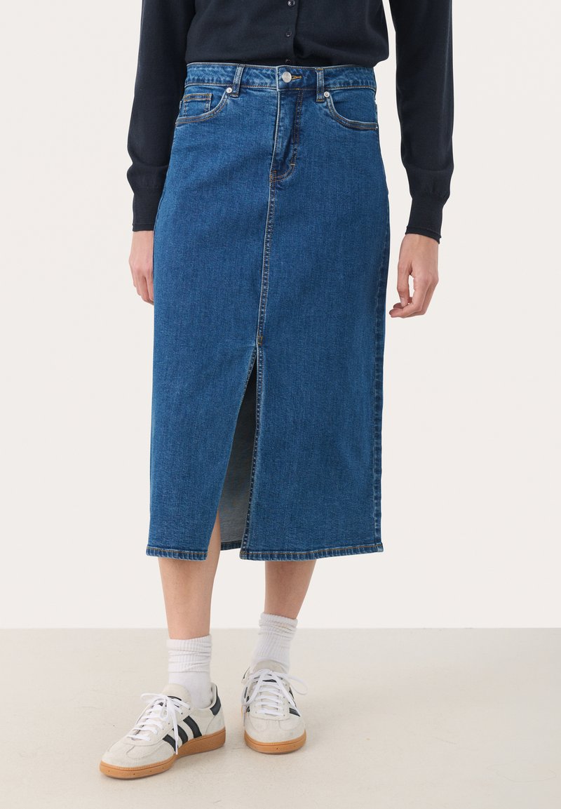 Denim svinčnik krilo v temno modri barvi z sprednjim razporkom, standardnim pasom in petimi žepi; oblečeno s športnimi belimi supergami in belimi nogavicami.