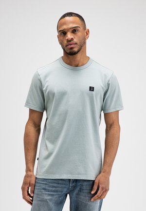 Man draagt een lichtgrijs T-shirt met korte mouwen en een blauwe spijkerbroek, staand tegen een effen witte achtergrond.
