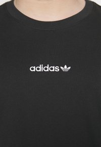 Czarny bawełniany t-shirt z okrągłym dekoltem oraz białym haftowanym logo "adidas" i symbolem trifolium na piersi. Gładka faktura.