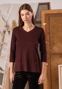 Bordowy sweter z dekoltem w serek i rękawami o długości do łokcia, wykonany z miękkiego materiału, z prostym dołem i małą złotą metką jako akcentem.