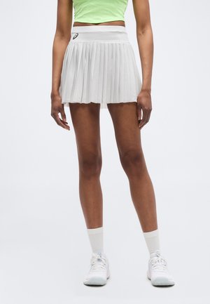 Femme portant une jupe de tennis plissée blanche, des chaussures de sport blanches et un haut court vert clair sur un fond blanc uni.