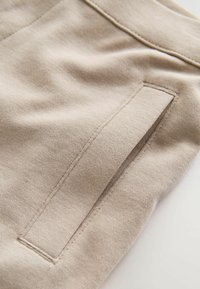 Zbliżenie na beżową tkaninę z naszytą kieszenią o czystych liniach i gładkiej teksturze. Miękki, lekki materiał z delikatnym połyskiem.