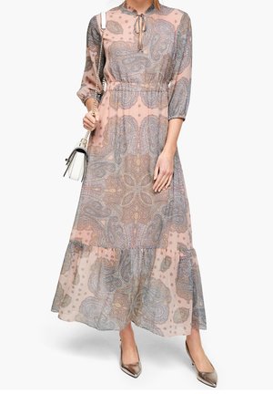 Kobieta ubrana w długą sukienkę o wzorze paisley w kolorze różowym z rękawami 3/4, trzymająca białą torebkę i nosząca metaliczne szpilki z ostrym noskiem.