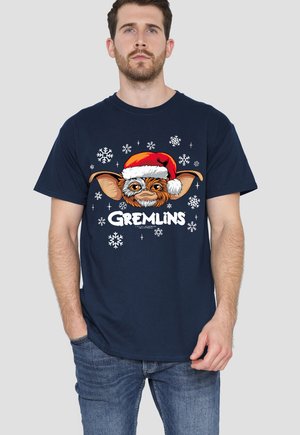 Mann trägt ein marineblaues T-Shirt mit einem Gremlins-Charakter, der einen Weihnachtsmannhut trägt, umgeben von Schneeflocken, kombiniert mit blauen Jeans.