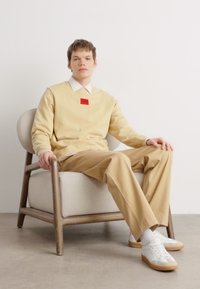 Felpa beige con un'etichetta rossa, abbinata a pantaloni beige chiaro. Modello seduto su una sedia color sabbia chiara con braccioli in legno, indossando sneaker bianche.