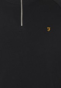 Tissu côtelé noir avec une fermeture éclair argentée en haut au centre et une petite lettre "F" jaune brodée sur le côté droit.