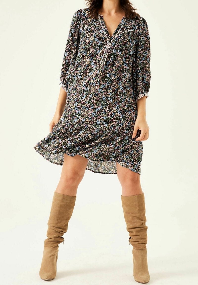 Femme portant une robe ample à motif floral avec manches 3/4 et des bottes en daim beige montant jusqu'aux genoux, debout devant un fond uni.