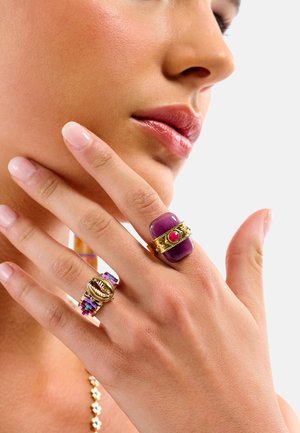 Bagues en or ornées d'une grosse pierre violette avec un accent rouge et un design décoratif, accompagnées d'une bague multicolore à perles avec un coquillage cauri.