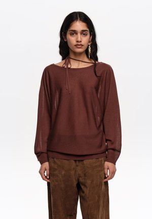 Ženska z dolgimi temnimi lasmi, oblečena v ohlapen rjav pulover, ujemajoče hlače in zlato viseče uhan, stoji na belem ozadju.