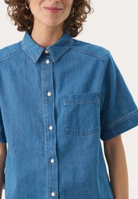 Camicia in denim a maniche corte di colore azzurro chiaro, con colletto a camicia, taschino frontale e dettagli di cucitura discreti. Tessuto dalla texture liscia.