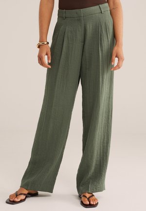 Žena nosi vysoké pásové široké nohavice z olivovo zelenej ľanovej látky s záhybmi, hnedé tielko, zlatý náramok a čierne sandále na béžovom pozadí.