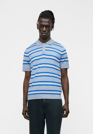 Hombre vistiendo una camisa polo de manga corta azul claro con rayas horizontales azules y blancas y pantalones negros, de pie frente a un fondo liso.