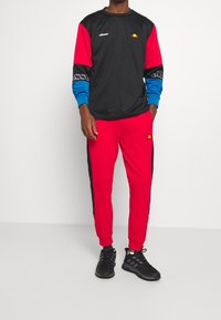 Person med svart, röd och blå långärmad sporttröja, matchande röda träningsoverallsbyxor och svarta löparskor, som står mot en vit vägg.