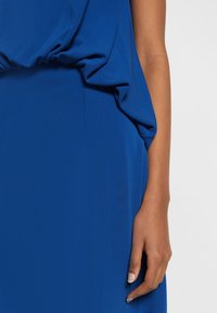 Bras droit et main d'une personne portant un vêtement bleu avec un tissu froncé près de la taille sur un fond uni.