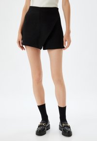 Shorts noirs en tweed taille haute avec un design asymétrique, texture lisse et coupe nette, associés à des mocassins noirs et des chaussettes.