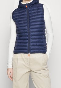 Gilet imbottito blu navy con cappuccio, design trapuntato, cerniera frontale con tiretto arancione; indossato sopra una maglia bianca a maniche lunghe.