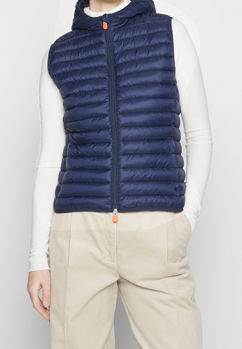 Gilet imbottito blu navy con cappuccio, design trapuntato, cerniera frontale con tiretto arancione; indossato sopra una maglia bianca a maniche lunghe.