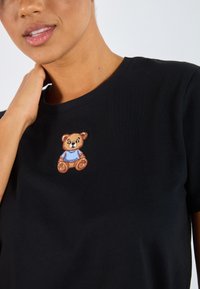Black cotton t-shirt featuring a stitched brown teddy bear with a blue shirt on the chest area. Short sleeves and a classic crew neckline.
