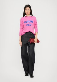 Frau in einem rosa Pullover mit der Aufschrift "ZUKÜNFTIGE CEO", dunklen weiten Hosen, schwarzen Schuhen, mit einer roten Clutch in der Hand, vor einem weißen Hintergrund stehend.