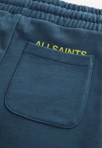 Granatowe dresy mają teksturowany materiał, elastyczny pasek w talii oraz tylną kieszeń z napisem "ALL SAINTS" w jaskrawym żółtym kolorze.