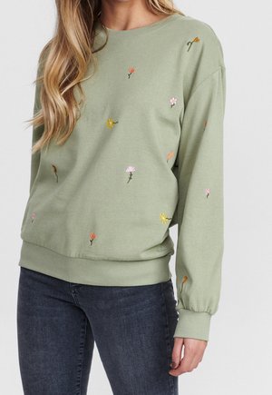 Femme portant un sweat-shirt vert clair avec de petites fleurs colorées brodées, associé à un jean bleu foncé.