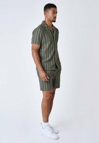 Camicetta verde oliva a maniche corte e pantaloni corti abbinati con righe bianche verticali, realizzati in un tessuto testurizzato. Abbinata a sneaker bianche.