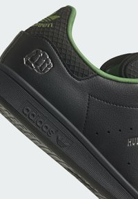 Czarny sneaker z teksturowaną skórzaną cholewką, zielonymi akcentami oraz wyraźnym tłoczonym motywem pięści. Posiada perforacje i logo po boku.