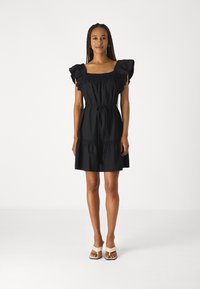 Rochie neagră din bumbac cu mâneci scurte volanate, decolteu pătrat, talie strânsă și fustă evazată, legată la talie. Modelul poartă sandale bej.