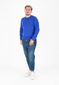 Niebieski sweter z okrągłym dekoltem wykonany z dzianiny, w połączeniu ze średnio zafarbowanymi dżinsami i białymi trampkami. Prosty fason z ściągaczami przy mankietach.
