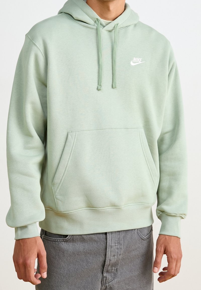 Homme portant un sweat à capuche Nike vert clair avec cordons et poche kangourou à l'avant, associé à un jean gris.