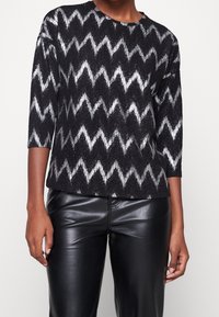 Svart och silver zigzag-mönstrad tröja med trekvartslånga ärmar, rund halsringning och ett mjukt, strukturerat tyg. Bärs tillsammans med svarta läderbyxor.