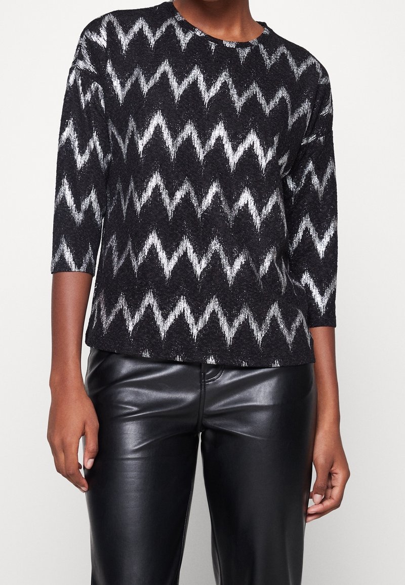 Svart och silver zigzag-mönstrad tröja med trekvartslånga ärmar, rund halsringning och ett mjukt, strukturerat tyg. Bärs tillsammans med svarta läderbyxor.