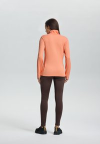Oranje textuur met lange mouwen top met een hoge kraag, gecombineerd met een paarse legging en zwarte schoenen met gele accenten.
