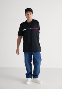 Crna majica kratkih rukava s bijelim i crvenim vodoravnim prugama, s logom "TOMMY HILFIGER". Uparena je s plavim trapericama i laganim tenisicama.