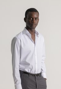 Camisa blanca de botones con cuello extendido, combinada con pantalones grises y un cinturón negro. Tela suave con mangas largas y un diseño ajustado.