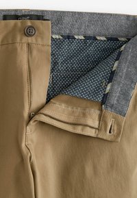 Pantalon marron avec une fermeture éclair et un bouton ouverts, montrant la doublure intérieure bleue à motifs et le détail gris de la ceinture.