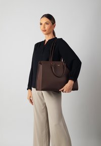Brown leather tote bag with textured surface, dual handles, and gold hardware accents. Paired with a black blouse and cream wide-leg trousers.