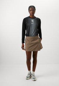 Long-sleeve black top with vertical stripes and logo, paired with a light brown knee-length skirt featuring button details. Grey sneakers complete the look.
