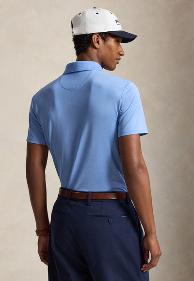 Uomo visto da dietro che indossa una polo azzurro chiaro, pantaloni blu scuro con cintura marrone, un cappellino da baseball bianco e blu scuro e un orologio da polso marrone.