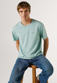 Mladý muž s krátkymi svetlohnedými vlasmi, oblečený v mentolovo zelenej tričku a modrých džínsoch, sediaci na drevenej stoličke proti jednému jednoliatemu pozadiu.