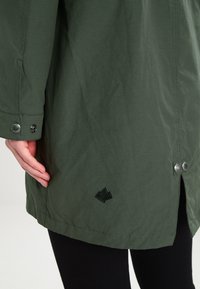 Grön, lätt jacka med en slät yta, tryckknappar och en broderad logotyp vid fållen. Har långa ärmar och rak skärning.