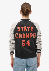 Sort og grå varsityjakke med "STATE CHAMPS 54" broderet i rødt. Den har ribstrikkede manchetter og kant, skulderlapper og en afslappet pasform.
