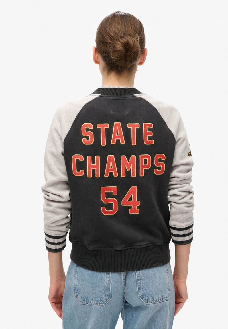 Sort og grå varsityjakke med "STATE CHAMPS 54" broderet i rødt. Den har ribstrikkede manchetter og kant, skulderlapper og en afslappet pasform.