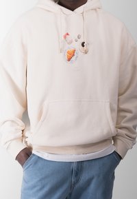 Beige hættetrøje med et madtematisk grafisk design, der inkluderer en croissant, kaffe og andre genstande. Lavet af blødt materiale, med en frontlomme og justerbar hætte.