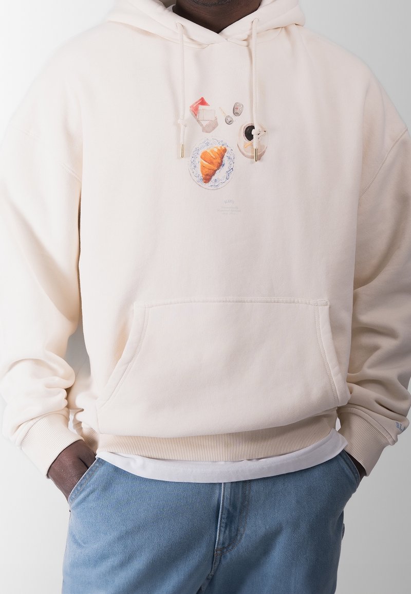 Beige hættetrøje med et madtematisk grafisk design, der inkluderer en croissant, kaffe og andre genstande. Lavet af blødt materiale, med en frontlomme og justerbar hætte.