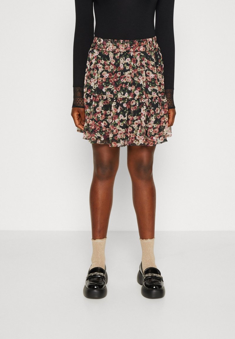 Femme portant un haut noir à manches longues, une mini-jupe fleurie avec des fleurs roses et blanches, des chaussettes beiges, et des mocassins noirs en cuir verni.