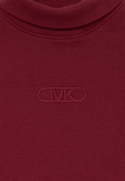 Close-up van een geribde bordeauxrode coltrui met een verhoogd MK-logo geborduurd op de borst.