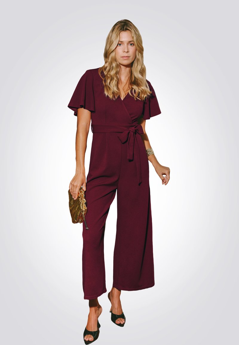 Bordó overal s krátkými křidélkovými rukávy, páskem v pase, širokými nohavicemi a texturovanou zlatou kabelkou. Na nohou jsou černé sandály s otevřenou špičkou.